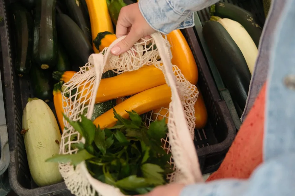 Cropped hands holding a cotton mesh bag of yellow zucchini and herbs at a vegetable market.
