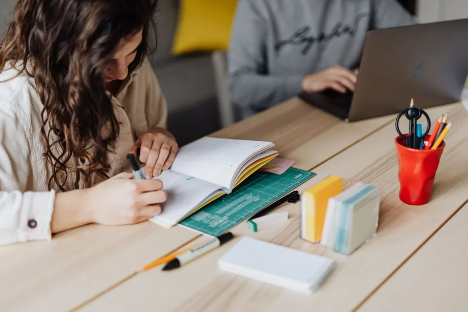 Woman writing in a paper planner at a table while another person works on a laptop nearby.